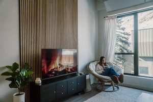 Wohnzimmer mit markanter WoodUpp-Lamellenwand aus naturfarbenen, dünnen, vertikal angeordneten Holzleisten hinter einem Fernseher, der ein Kaminfeuer zeigt, schwarzem TV-Sideboard, grüner Zimmerpflanze und einer runden Papasansessel mit Frau, die am großen Fenster entspannt ein Buch liest, mit bequemen Kissen, hellem Teppich und einer Duftkerze auf dem Sideboard im lichtdurchfluteten Raum.