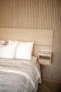 Schlafzimmeraufnahme mit WoodUpp-Vertikal-Lamellenwand in hellem Holz, sichtbarer Maserung und schmalen Schattenfugen, dazu ein passendes WoodUpp-Kopfteil aus Eiche. Rechts hängt ein schwebender WoodUpp-Nachttisch mit Marmorplatte, offenem Fach und Schublade. Weiße Bettwäsche und strukturierte Kissen betonen die warme, minimalistische Atmosphäre auf hellem Fischgrätparkett.