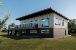 Modernes zweistöckiges Haus mit dunklem Klinkersockel, großen Panoramafenstern und überstehendem Flachdach. Obere Etage ist mit WoodUpp vertikalen Holzlamellen als Fassadenverkleidung versehen; WoodUpp-Holzverkleidung akzentuiert auch den Balkonbereich. Glas-Balkongeländer, gepflegte Rasenfläche und Bäume im Hintergrund, in warmem Naturton, schlanke, gleichmäßige Lamellen und klare Linien betonen die moderne Architektur.