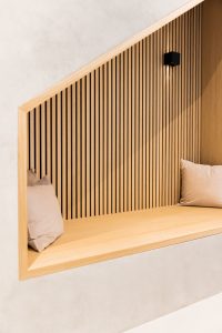 WoodUpp: Eingelassene Sitznische aus hellem Holz mit senkrechten WoodUpp-Lamellenpaneelen, feiner Rahmenkonstruktion und eingelassener Sitzbank aus Eichenholz. Zwei bequeme beige Kissen und eine kleine schwarze Wandleuchte ergänzen das warme, minimalistische Ambiente; dezente Schattenfugen, abgeschrägte Rahmenkante und sichtbare Holzmaserung betonen die Optik.