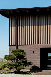 WoodUpp trælameller som lodret slat-facade dækker øverste etage: tætte, lyse, naturlige træstrimler i jævne mellemrum, monteret som facadebeklædning over mørk teglvæg. Billedet viser WoodUpps vertikale facadeprofiler tydeligt, og lamellerne har varme trætoner, slanke profiler og danner skarpe skygger i sollys.