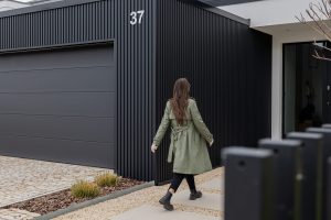 Kvinde i grøn trenchcoat går mod et moderne hus med WoodUpp sort, lodret trælamelfacade i antracit ved garage og indgang, samt WoodUpp sorte lamelhegn i forgrunden. Billedet viser også garageport med horisontale felter, husnummer 37, stenbelægning, grussti og minimalistisk beplantning.